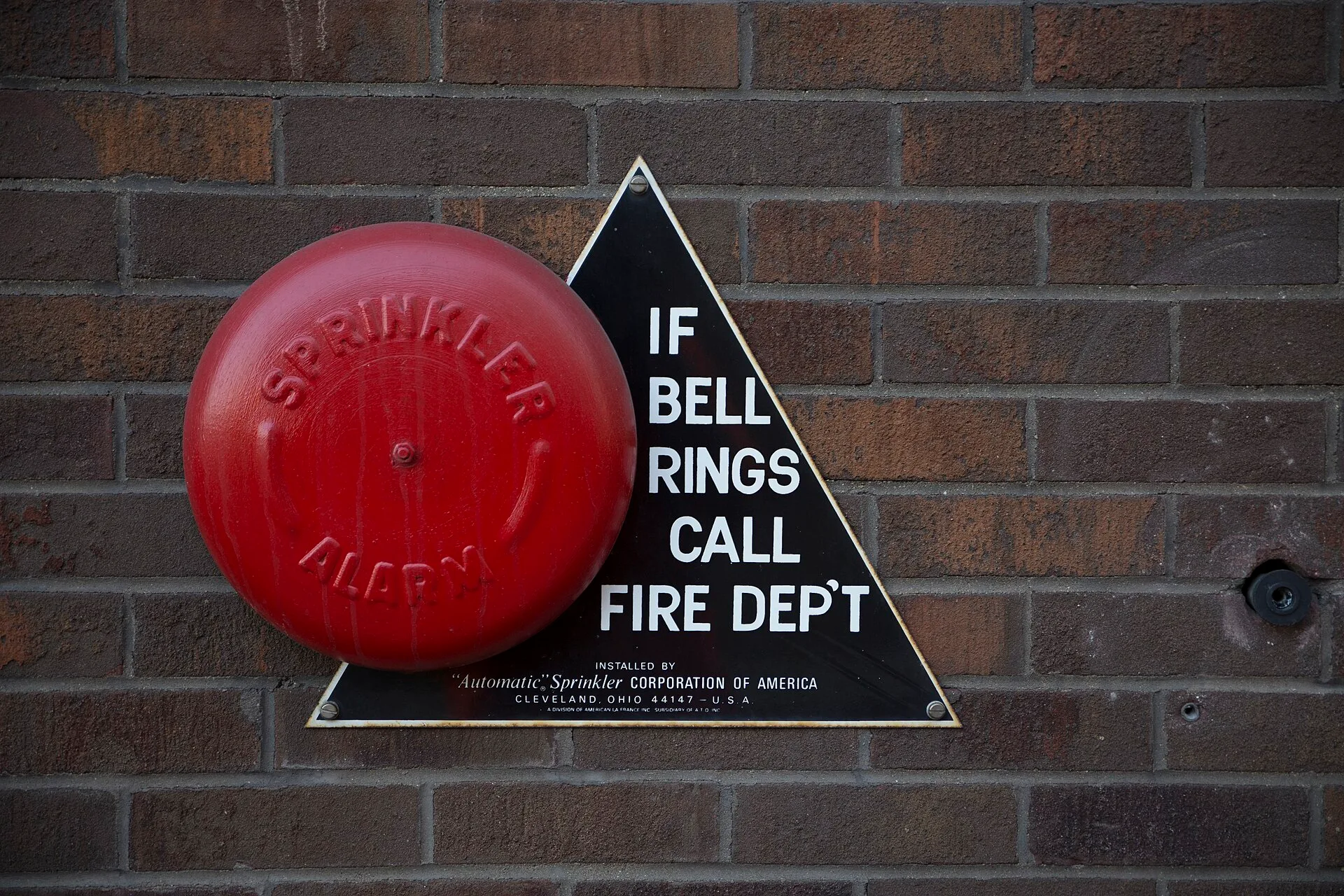 Red sprinkler alarm bell with IF BELL RINGS CALL FIRE DEPT sign on a brick wall