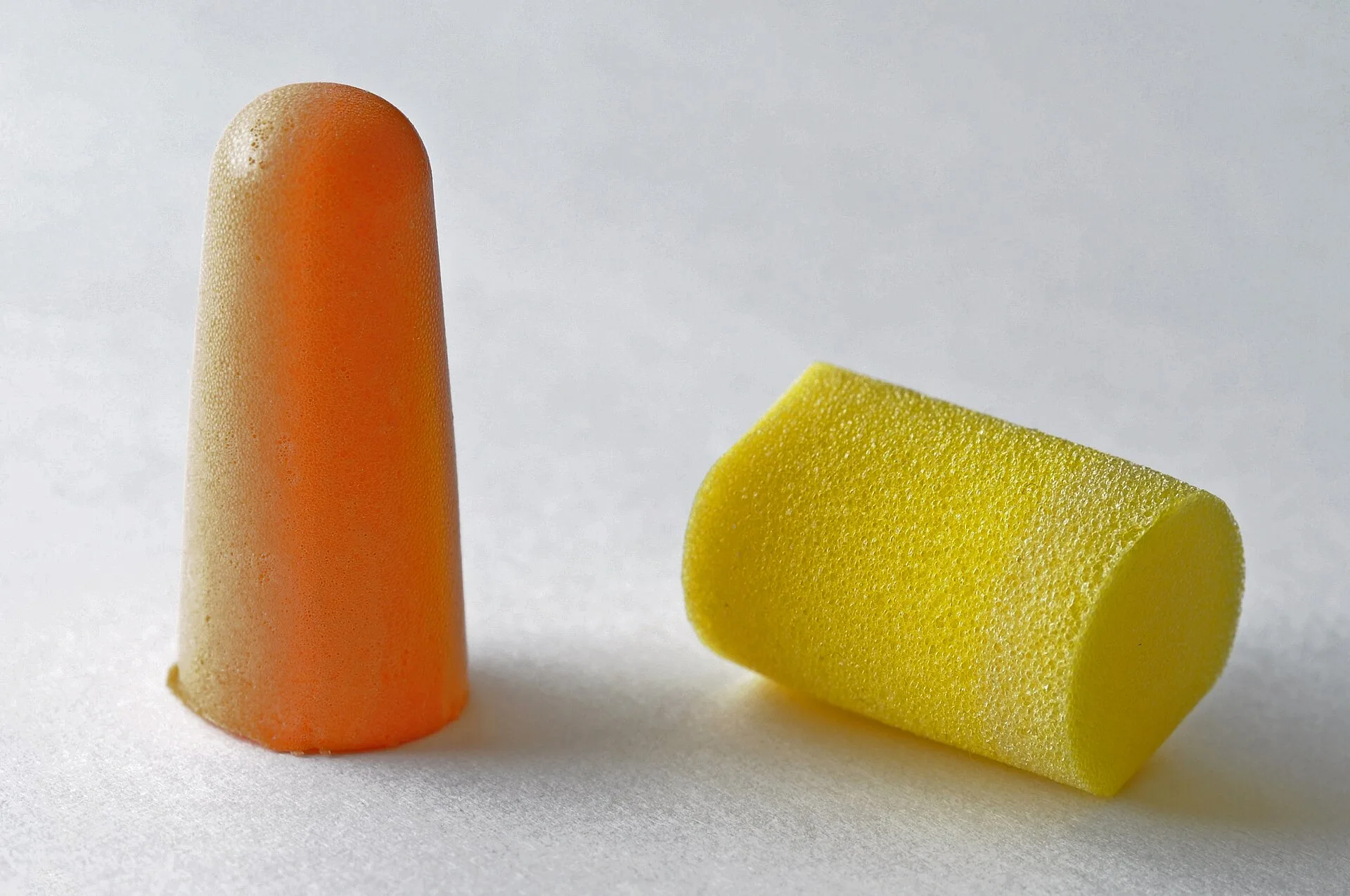 Orange tapered and yellow cylindrical foam earplugs on a light grey surface