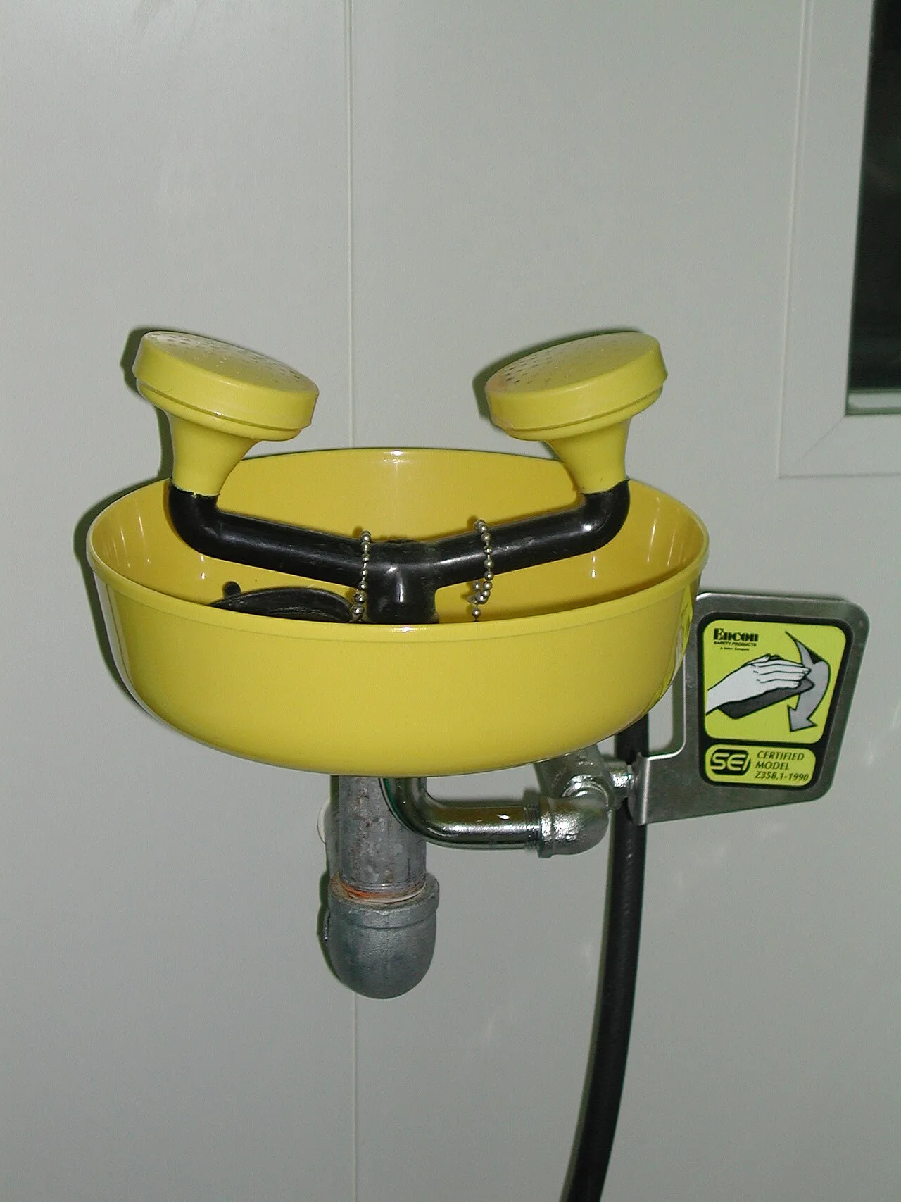 Wall-mounted plumbed eyewash station with twin spray heads, yellow bowl and push-flag activation