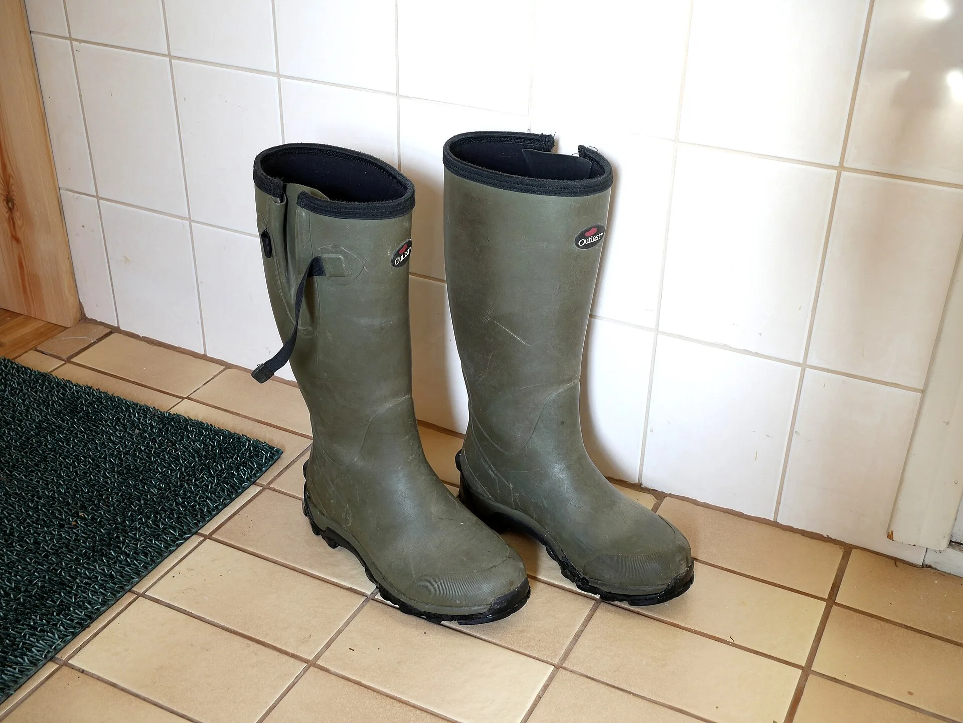 Pair of green rubber wellington boots standing by a tiled wall