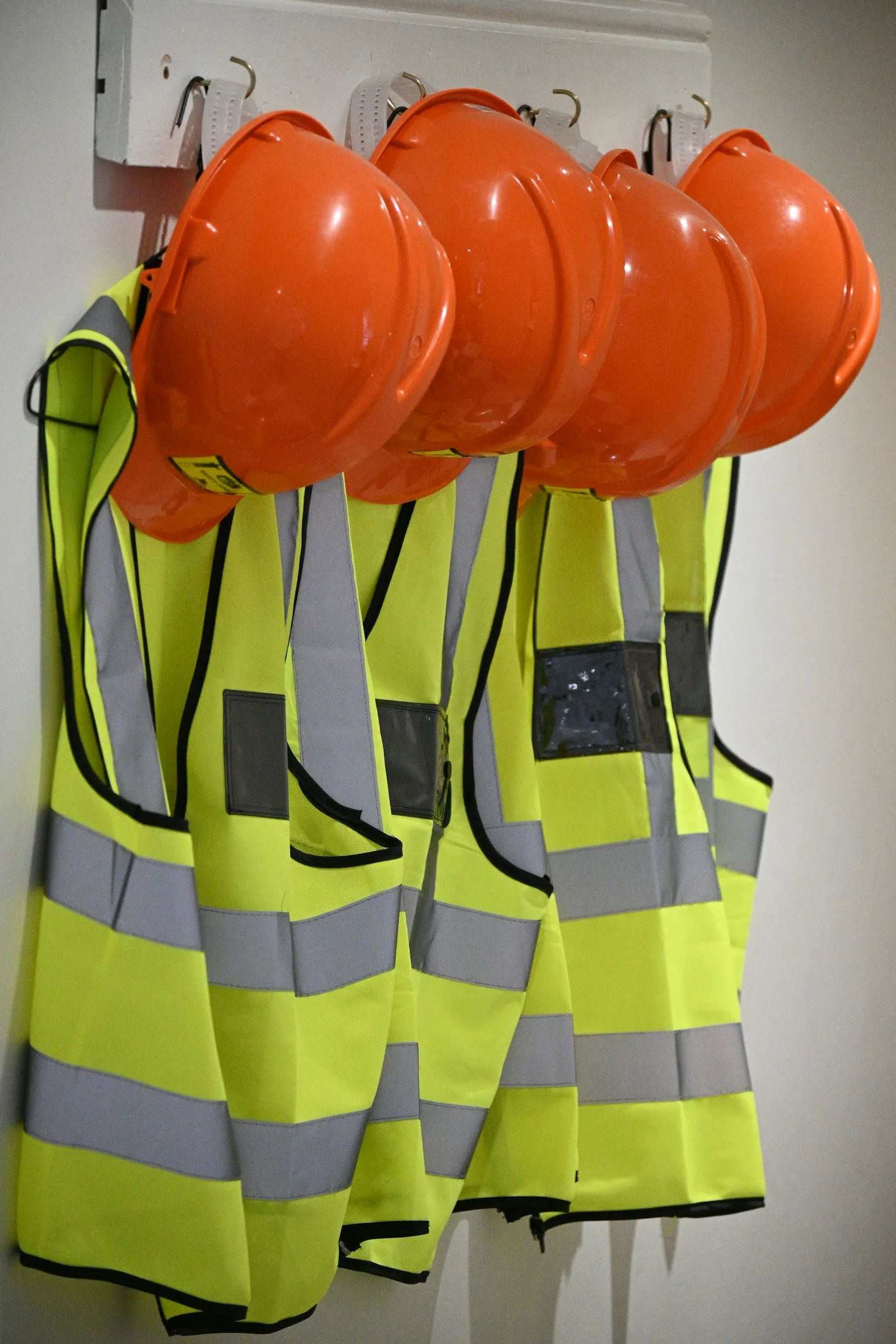 Four yellow high-visibility vests and orange hard hats hanging on wall hooks