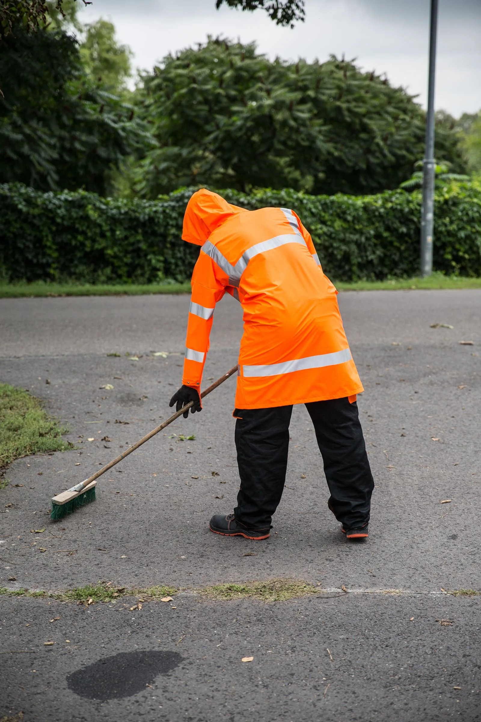 Street worker in an orange hooded high-visibility jacket with reflective tape sweeping a road
