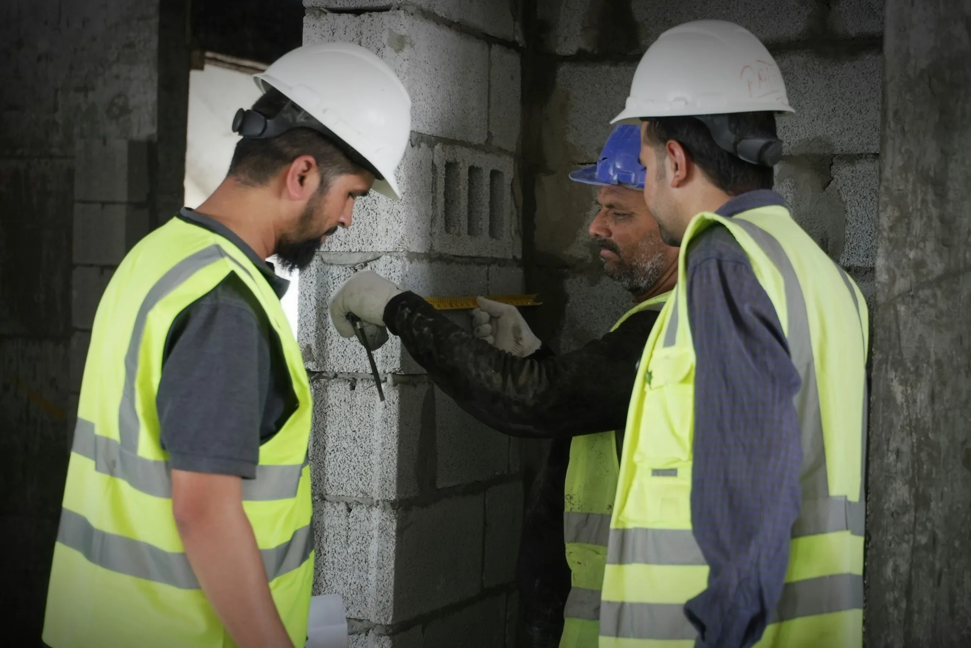 Three workers in hard hats and high-visibility vests measuring a concrete block wall on site