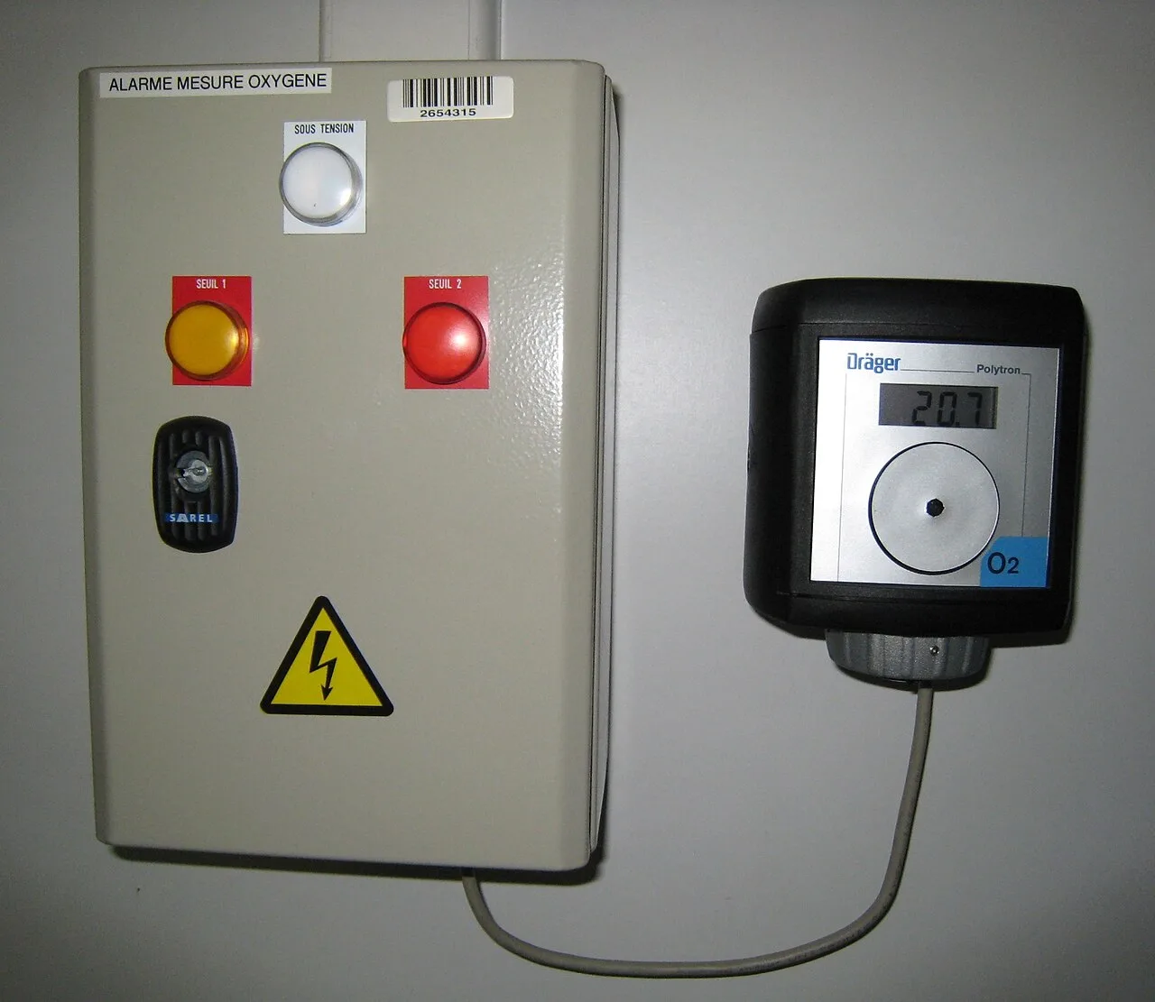 Wall-mounted fixed oxygen gas detector with digital display wired to an alarm control box with beacons
