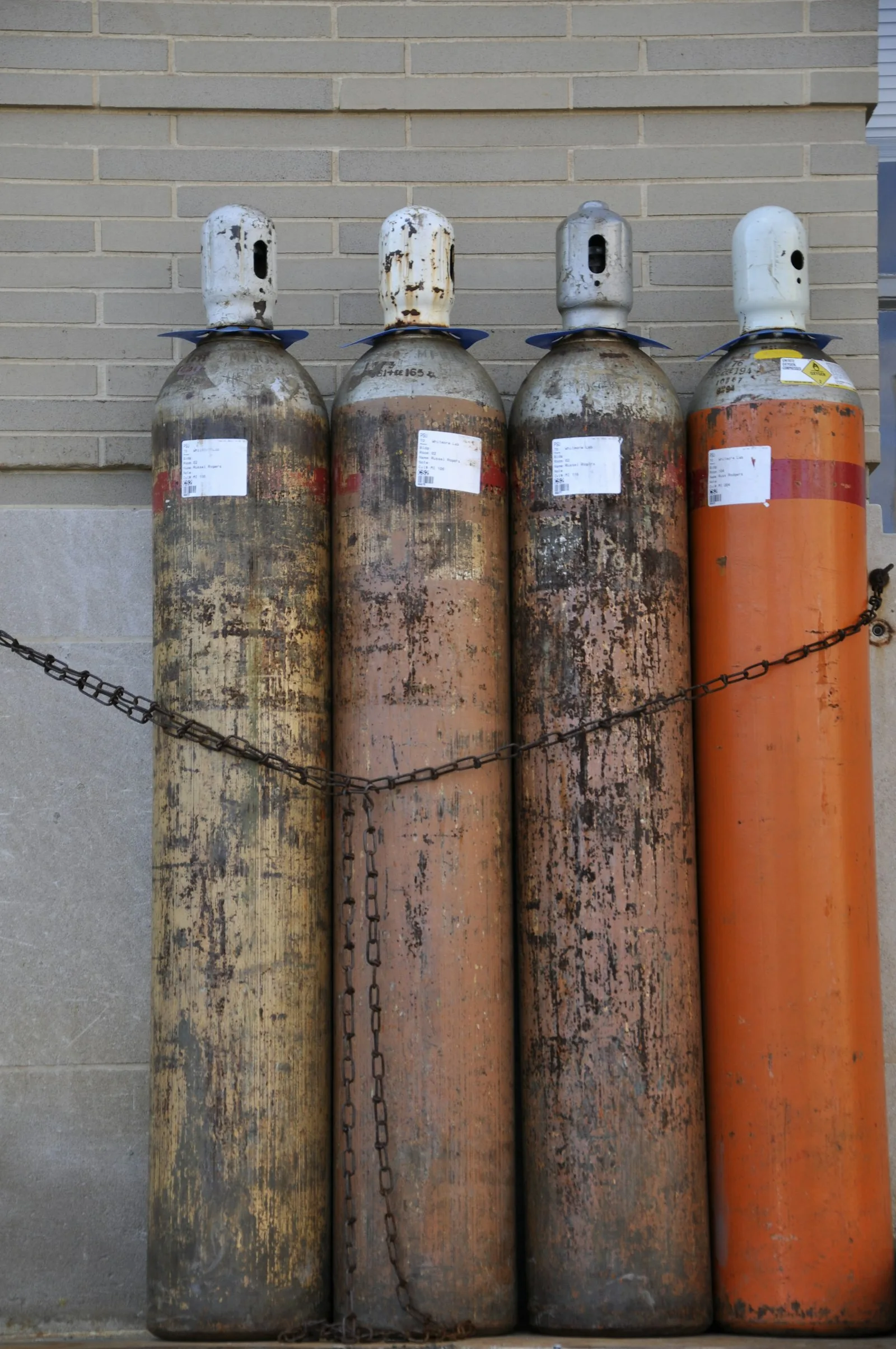 Four tall compressed gas cylinders secured upright with a chain against a wall